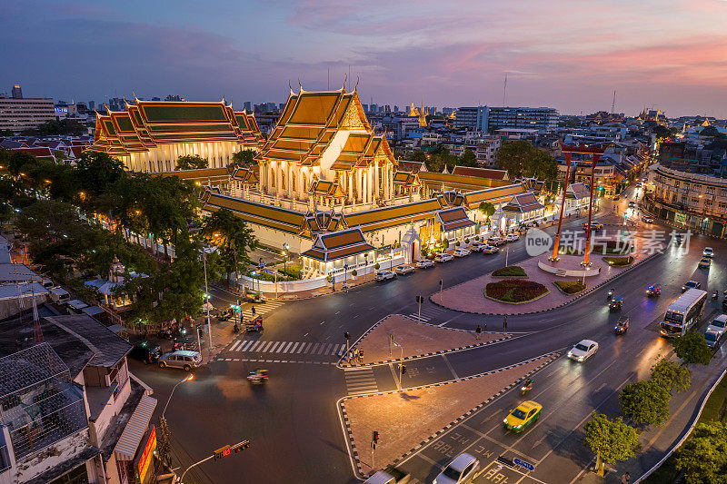 从空中无人机的角度看Suthat Thepwararam(Suthat Thepwararam寺)和巨大的秋千(Sao Chingcha)泰国曼谷，一个著名的旅游地标，黄昏时分车辆和游客沿着街道通勤。曼谷泰国